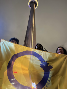 Three members of Intersex Canada holding up the organizations yellow and purple flag standing in front of the CN tower
